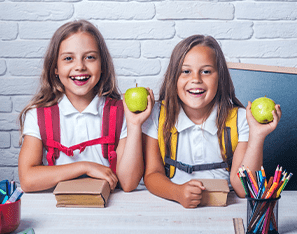 two students holding apples and sitting with backpacks on