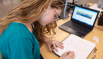 Student writing in a notebook next to a laptop
